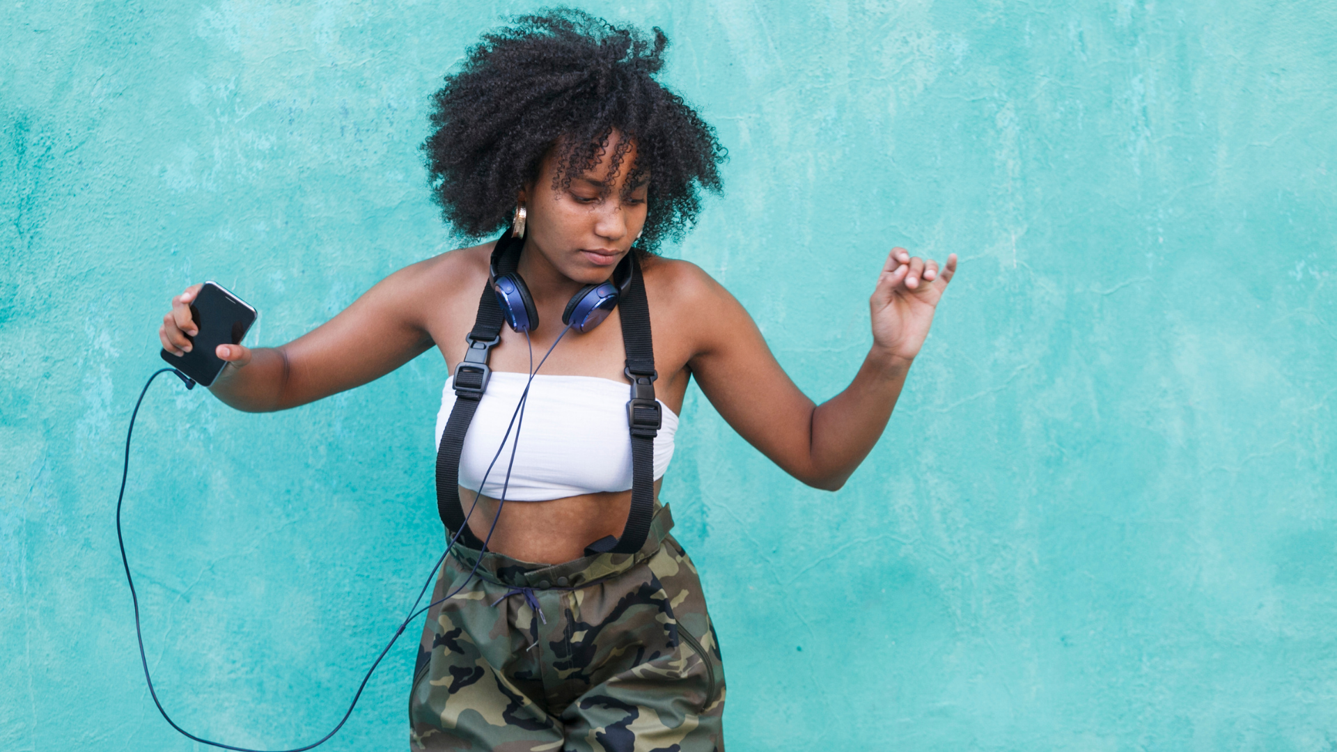 woman wearing camo pants and white crop top and headphones dancing against blue wall